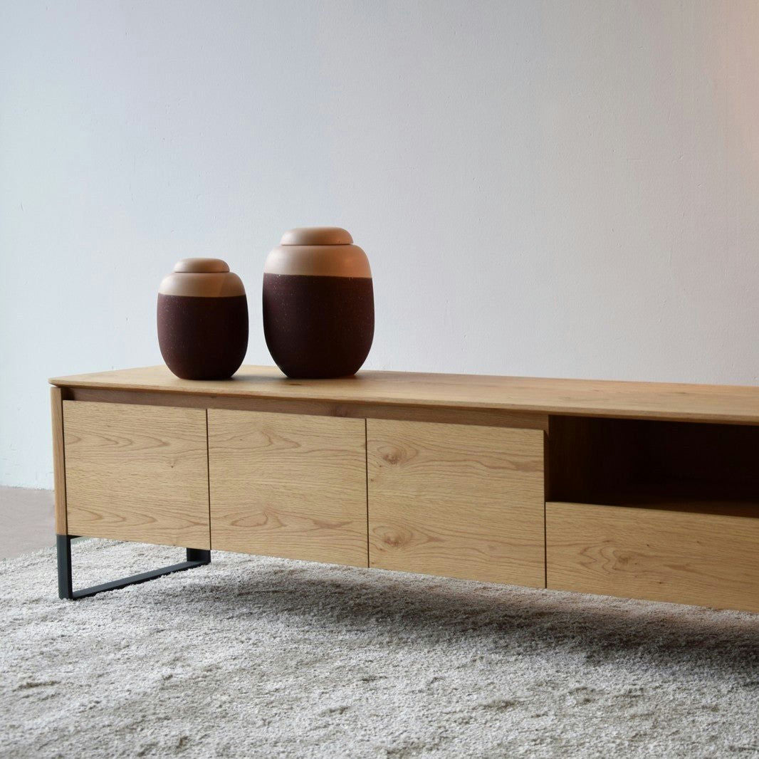 Un Mueble TV Roble Natural Mate y Grafito - Lignar con elegantes patas de metal negro ofrece amplio espacio de almacenamiento y sostiene dos jarrones de cerámica marrón en su superficie, que descansan elegantemente sobre una alfombra beige texturizada.