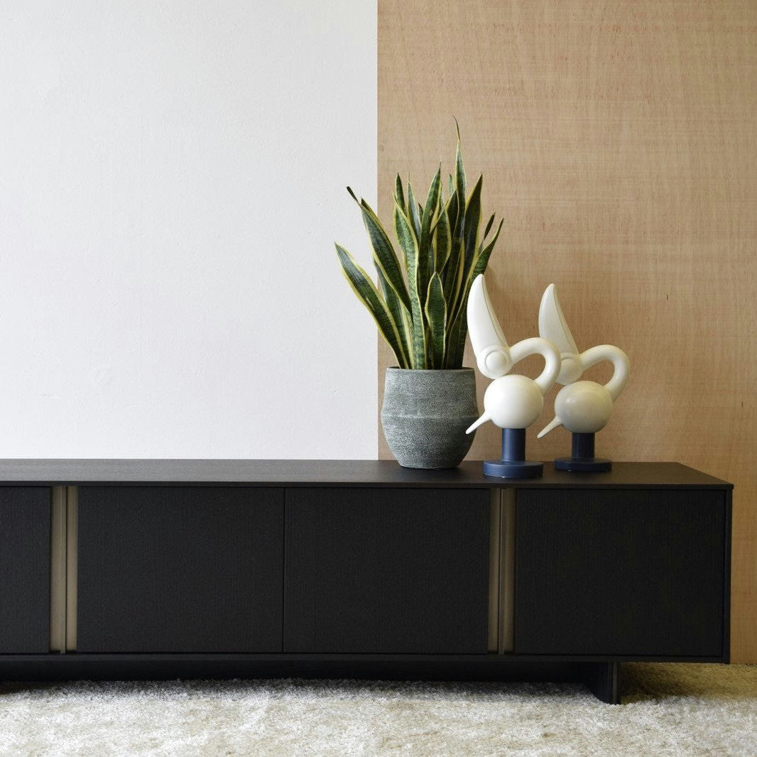 Una planta en maceta y dos esculturas abstractas adornan el Mueble TV Roble Negro Mate, Detalles Oro Viejo, contra una pared que contrasta. La escena se equilibra con una alfombra de color claro debajo, que realza la estética elegante.