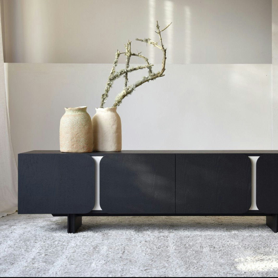 El Mueble TV Roble Negro mate Y Arena - Curva se encuentra sobre una alfombra texturizada, presentando un diseño moderno oscuro con bordes redondeados. Dos jarrones, incluido uno con una rama, se encuentran en la parte superior, realzando su elegante estética curva y encarnando el diseño contemporáneo.