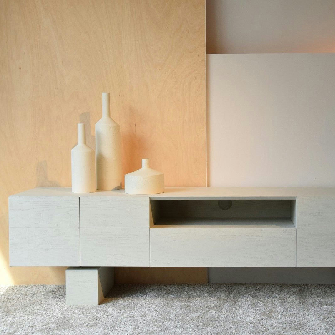 Un mueble TV minimalista Roble en Tono Blanco roto Mate con jarrones geométricos en la parte superior se presenta elegantemente sobre un fondo de paneles de madera, todo ello apoyado sobre una alfombra texturizada.