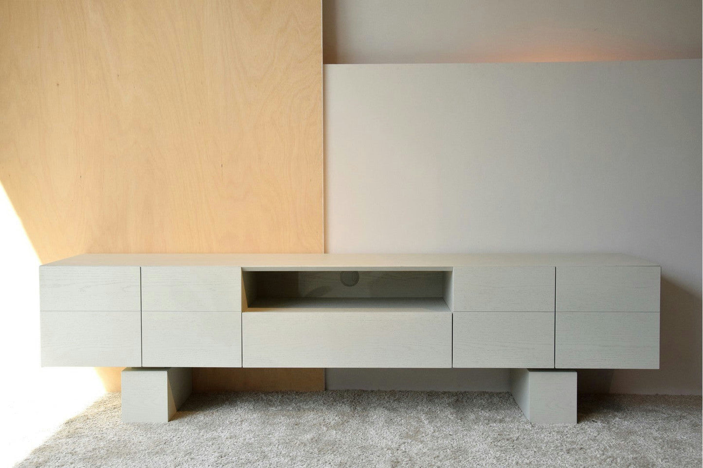El Mueble TV Roble en Tono Blanco Roto Mate es un aparador minimalista de madera de color claro con un diseño geométrico, que cuenta con seis cajones y un estante central abierto. Su elegante acabado complementa a la perfección la alfombra clara que se encuentra debajo.