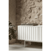 Un moderno mueble blanco con puertas nervadas junto a una pared de piedra, diseñado como Mueble TV Sierra 3P, con una planta a la izquierda y un tocadiscos encima.