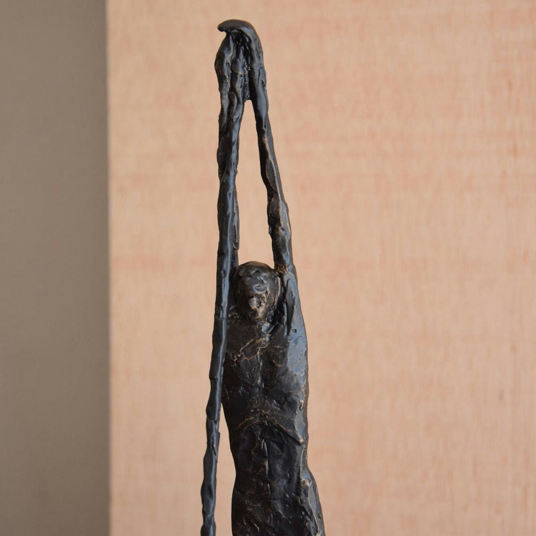 La Figura decorativa de metal en bronce envejecido N2 es una esbelta escultura humana que se extiende hacia arriba con ambos brazos, sobre un fondo neutro y con un acabado de bronce envejecido que agrega elegancia atemporal a cualquier diseño interior.