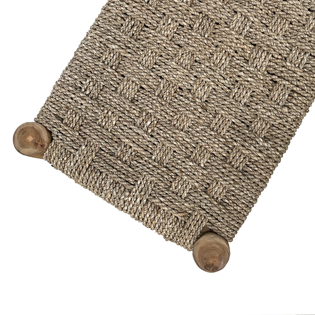 Primer plano de la Banqueta de Teca y Ratán Natura: cuenta con un asiento de ratán tejido con patrón a cuadros y patas de madera redondeadas, una pieza elegante que agrega estilo natural a cualquier espacio.