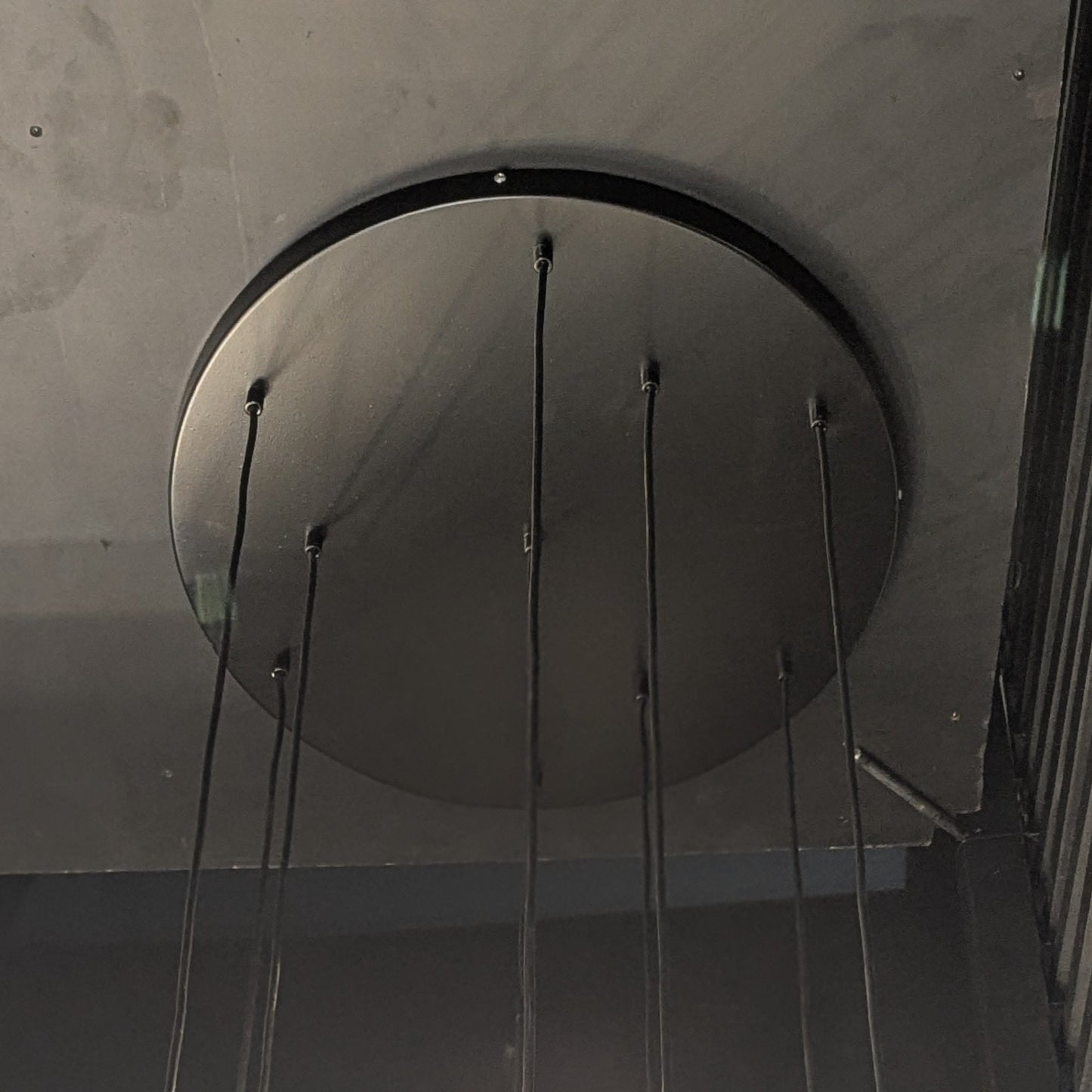 La Lámpara de techo en acero negro Candil Kairo 7 presenta un diseño circular negro con múltiples cables, que resalta su elegante elegancia sobre un fondo oscuro.