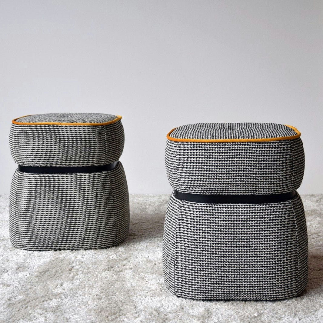 Dos pufs grises texturizados de la colección Pack 2 Puffs Cuadrados Tapizados Classic presentan franjas negras y detalles en amarillo, bellamente colocados sobre una alfombra clara contra una pared sencilla. Estos pufs combinan estilo y durabilidad con su diseño clásico y tapizado de poliéster.