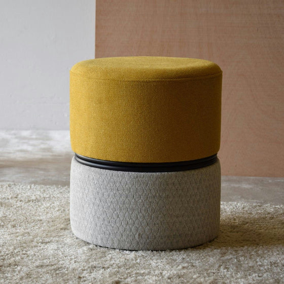 Un puf amarillo y gris de la colección Pack 2 Puffs Redondos Tapizados Yellkin se encuentra sobre una alfombra beige contra una pared marrón claro, encarnando el diseño contemporáneo.