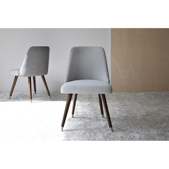 Dos elegantes sillas grises con patas de madera del Pack de 2 Sillas de Comedor Gris y Madera - Comfino están colocadas sobre un suelo alfombrado de color claro. Una silla está en primer plano mirando hacia adelante, mientras que la otra está al fondo ligeramente girada hacia un lado. Sillas de comedor perfectas para cualquier hogar contemporáneo.