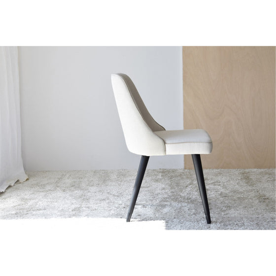 Vista lateral de una silla tapizada en color beige con patas de madera negra sobre una alfombra gris claro en una habitación minimalista con una pared blanca y de madera clara. Parte del elegante Pack de 2 Sillas de Comedor en Tonos Beige - Comodante, esta pieza agrega comodidad sofisticada a cualquier espacio de comedor.