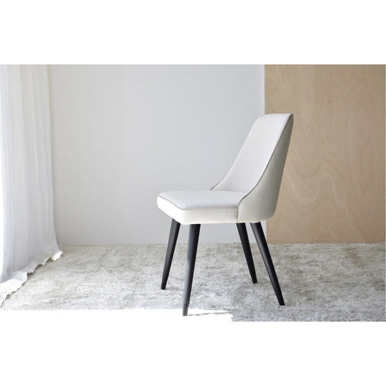 Una silla moderna con asiento y respaldo tapizados en blanco y patas de madera negras está colocada sobre una alfombra gris claro frente a una cortina blanca y un panel de pared de madera. Esta elegante pieza forma parte del Pack de 2 Sillas de Comedor en Tonos Beige - Comodante, perfecto para espacios de comedor contemporáneos.