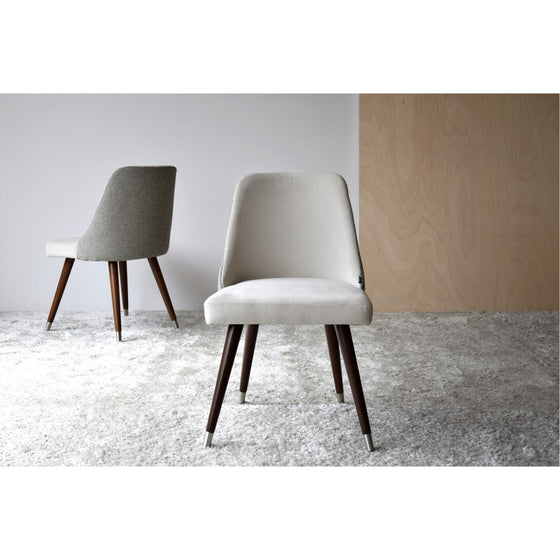 Dos modernas Sillas de Comedor en Tono Piedra y Gris - Piedrizzo Pack 2 tapizadas con patas de madera se colocan sobre una alfombra texturizada de color claro sobre un fondo liso y claro. Una silla mira hacia adelante mientras que la otra está inclinada hacia un lado, mostrando su tapicería de poliéster que combina comodidad y estilo a la perfección.