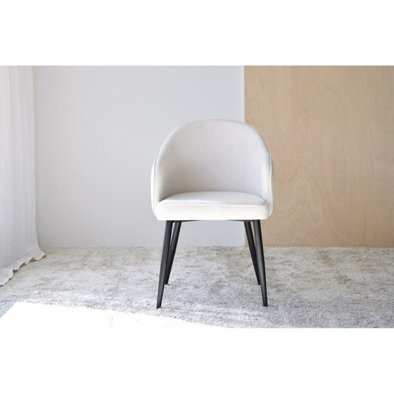 Un moderno Pack 2 Sillas de Comedor Tapizadas Roma tapizado en blanco con patas de metal negro se asienta sobre una alfombra texturizada de color claro sobre un fondo liso, ofreciendo un diseño sinuoso que combina a la perfección estilo y funcionalidad.