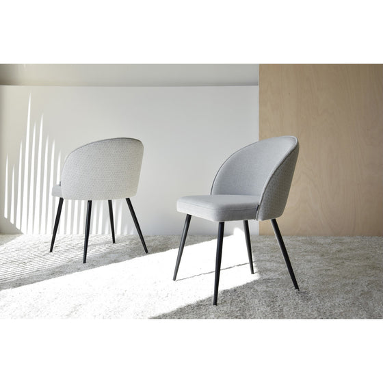 Dos modernas Sillas de Comedor Tapìzadas Sofistica Pack 2 de color gris con respaldo curvo y patas de metal negro se colocan sobre una alfombra de color claro en una habitación minimalista con paredes blancas y de madera. Las sombras de las persianas son visibles, realzando el diseño moderno.