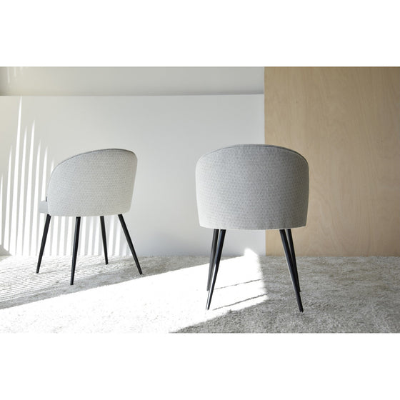 Dos modernas sillas de comedor tapizadas en blanco con patas negras miran hacia una pared de color claro en una habitación minimalista. La luz natural proyecta sombras en el suelo, realzando el elegante diseño moderno de este Pack 2 Sillas de Comedor Tapìzadas Sofistica.