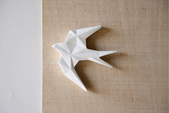 La Figuras decorativas en cerámica Blanco brillo Anduriña, una escultura de pájaro estilo origami blanco, está elegantemente montada en una pared con textura beige al lado de una elegante superficie vertical blanca, incorporando el estilo moderno y agregando sofisticación a su espacio.