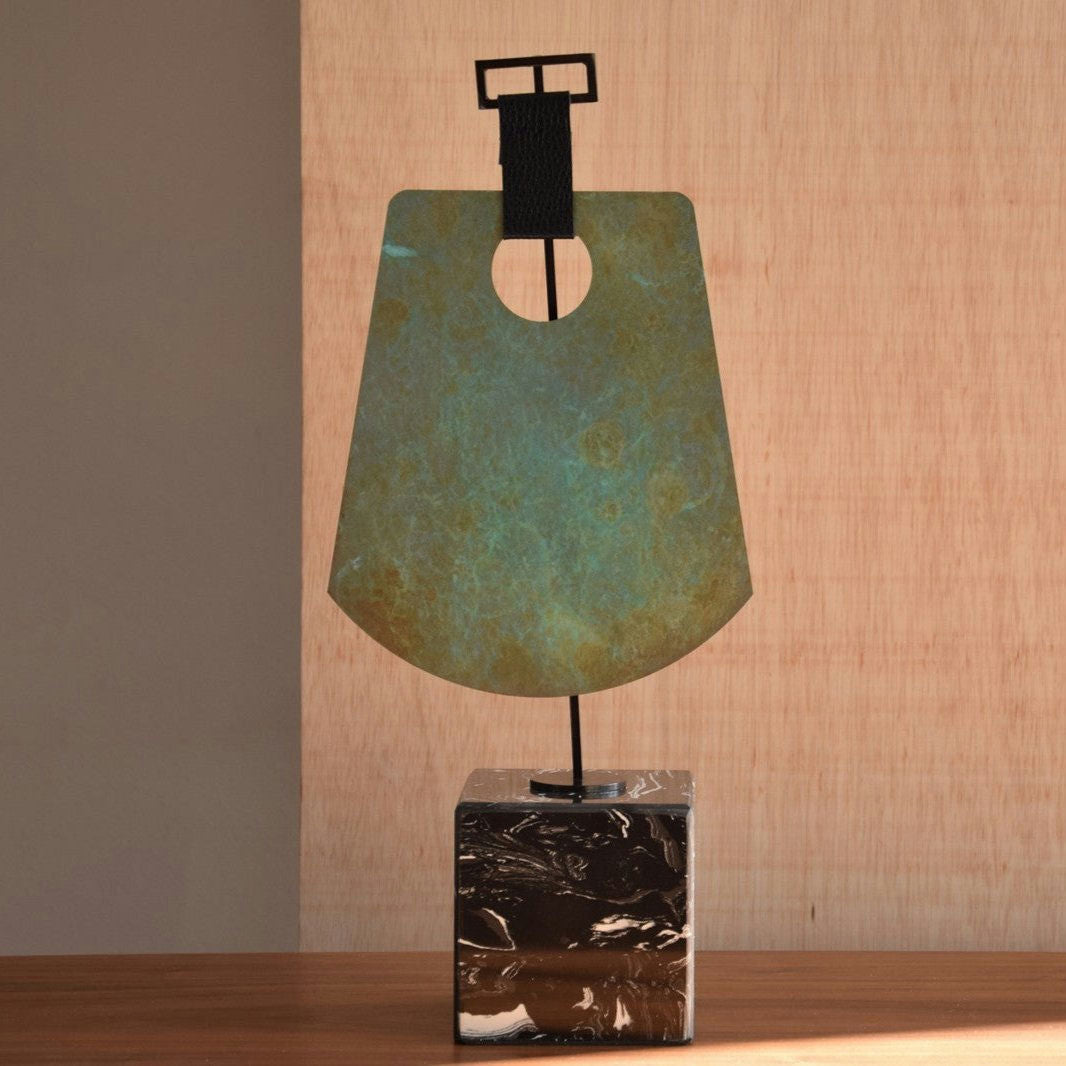 La Pieza Decorativa Etnica Acero-Mármol N1 presenta una escultura abstracta de metal con acabado pátina, con una parte superior rectangular y una parte inferior curva, elegantemente montada sobre una elegante base de mármol negro, perfecta contra un fondo de madera.