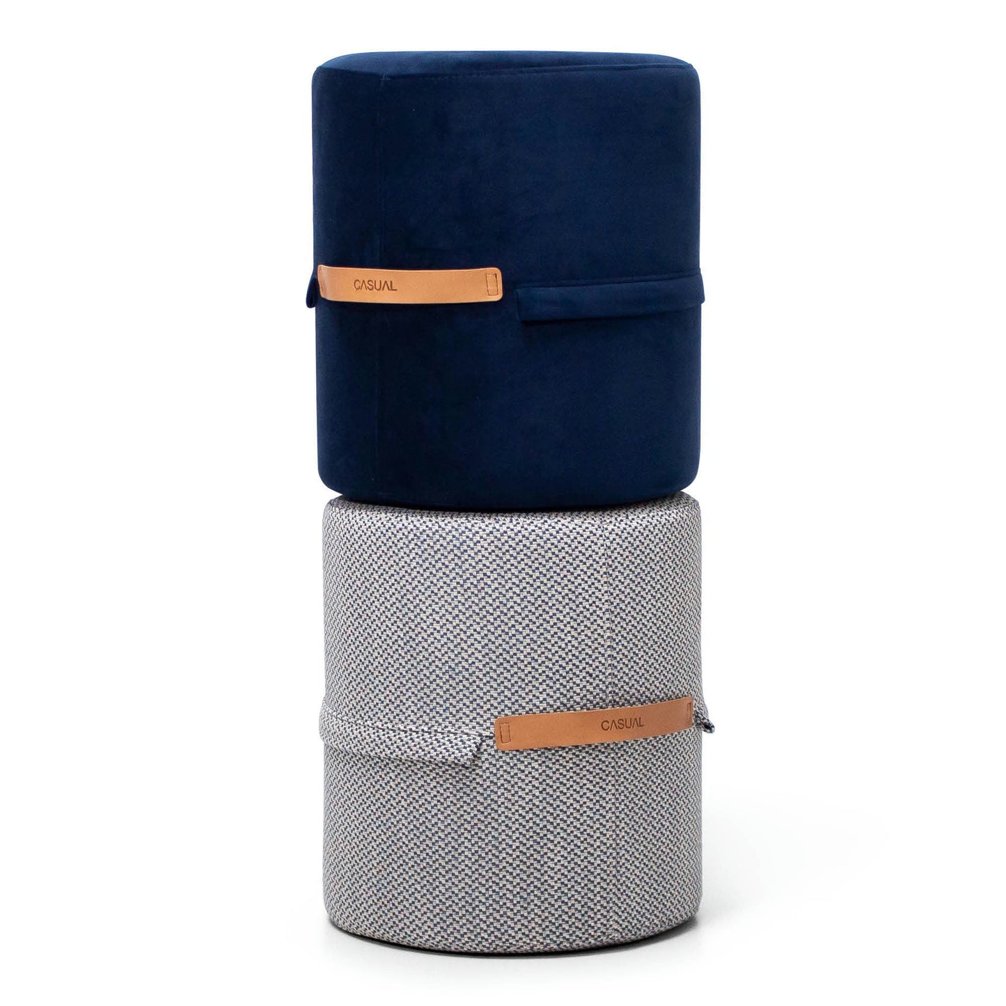 Dos otomanas apiladas: la superior es un Puff Redondo Tapizado Pull de terciopelo azul oscuro, la inferior es de espiga gris, ambas con un cinturón decorativo de cuero con la etiqueta "CASUAL", que personifica el diseño minimalista.