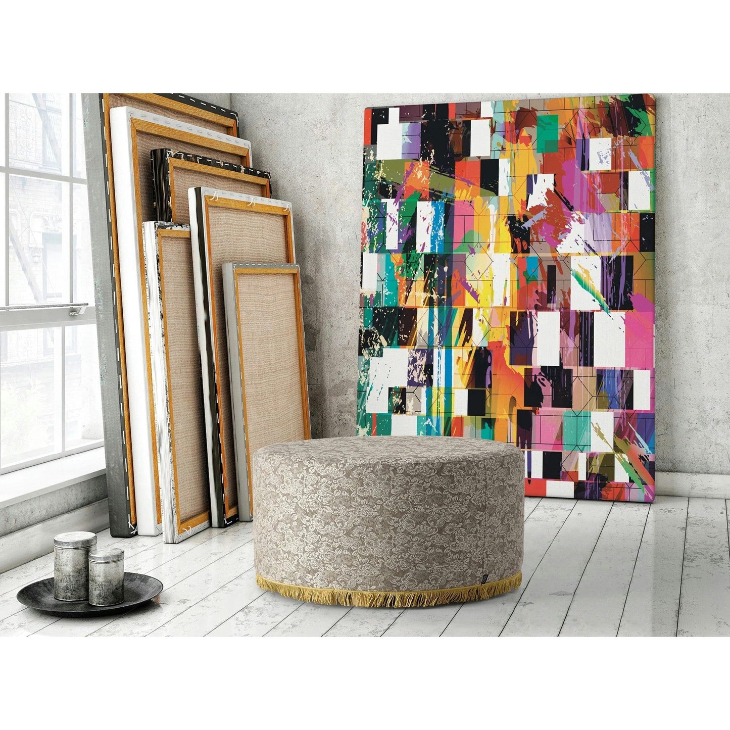 Una pintura abstracta colorida se apoya contra una pared con lienzos en blanco. Una otomana redonda Puff Redondo Tapizado reposa sobre un piso de madera blanca, mostrando una bandeja con tres objetos cilíndricos, capturando la esencia refinada del mobiliario contract.