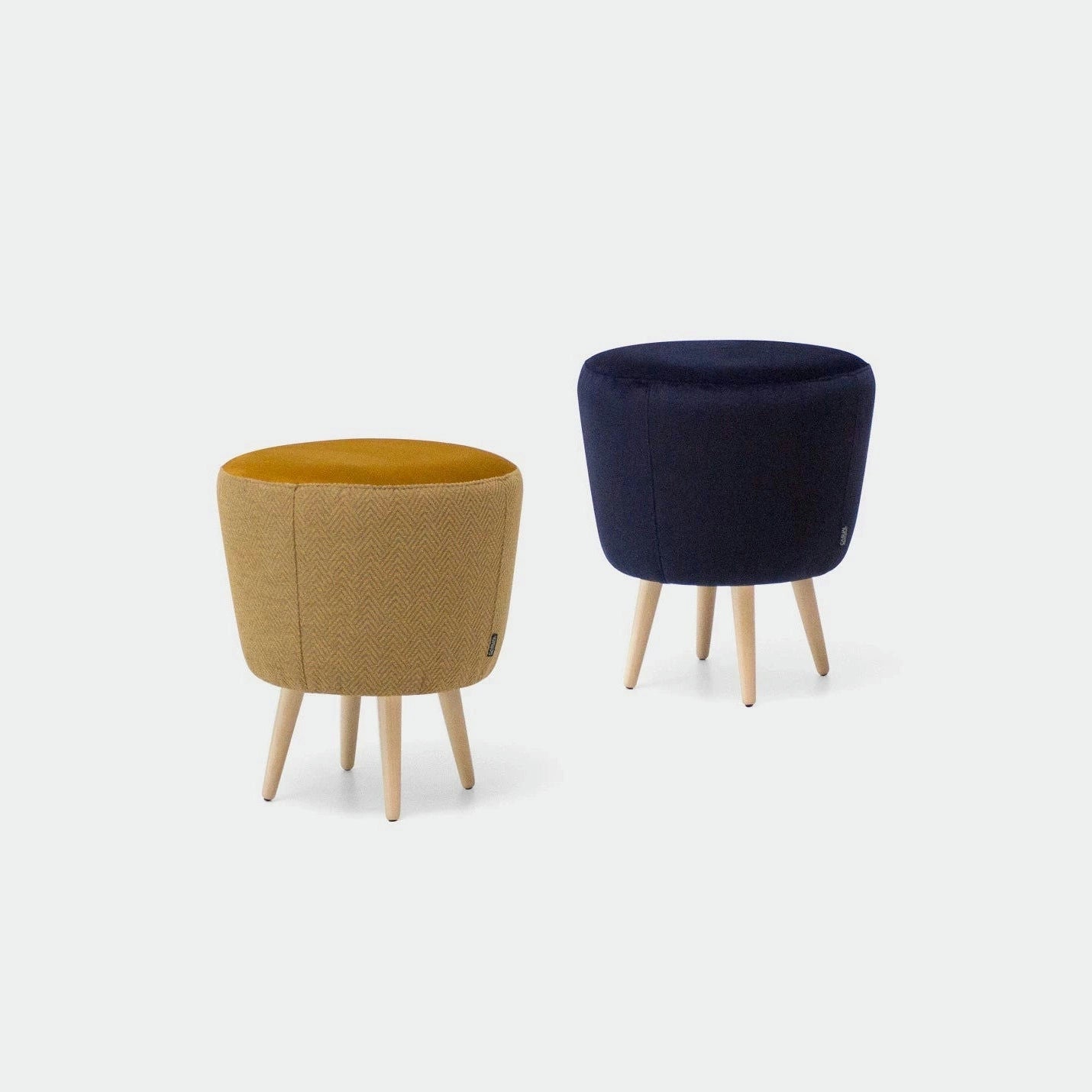 El Puff Redondo Tapizado con patas Jakoff consta de dos taburetes redondos con patas de madera de estilo nórdico; uno con asiento en color amarillo mostaza y el otro en color azul marino. Sobre un fondo claro, combinan estilo y funcionalidad sin esfuerzo.