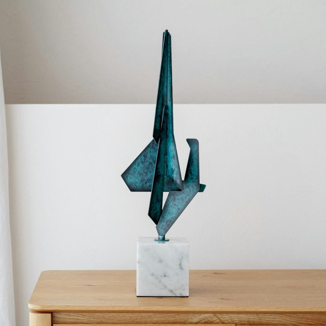 Figura Decorativa en Metal Verde Abstracción N1 presenta una escultura geométrica de metal sobre una base de mármol blanco, resaltando el diseño moderno. Colocado sobre una superficie de madera contra una pared clara.