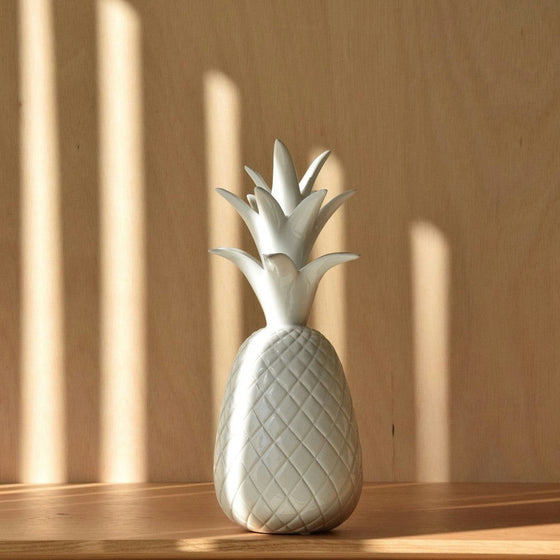 Las "Figuras Decorativas en Cerámica Blanca Piña" de la colección PIÑA adornan elegantemente una superficie de madera, proyectando intrincados patrones de luces y sombras en el fondo con su exquisito diseño artesanal.