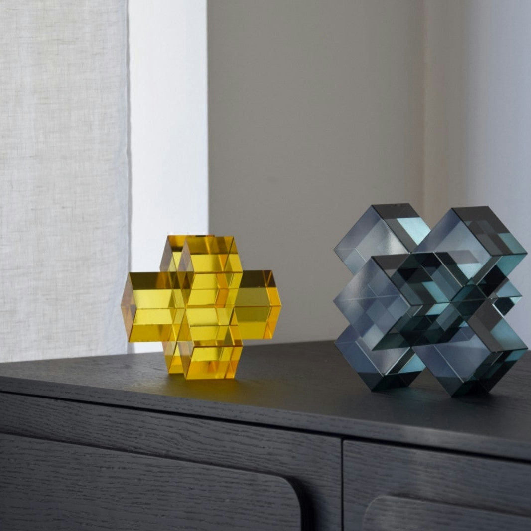 Una escultura geométrica amarilla y otra azul grisácea del "Set de 2 Piezas Cristal Color. Cubos" descansan sobre una superficie de madera oscura, mostrando diseños intrincados. Una cortina blanca en el fondo agrega elegancia a este conjunto de exquisitas esculturas decorativas.