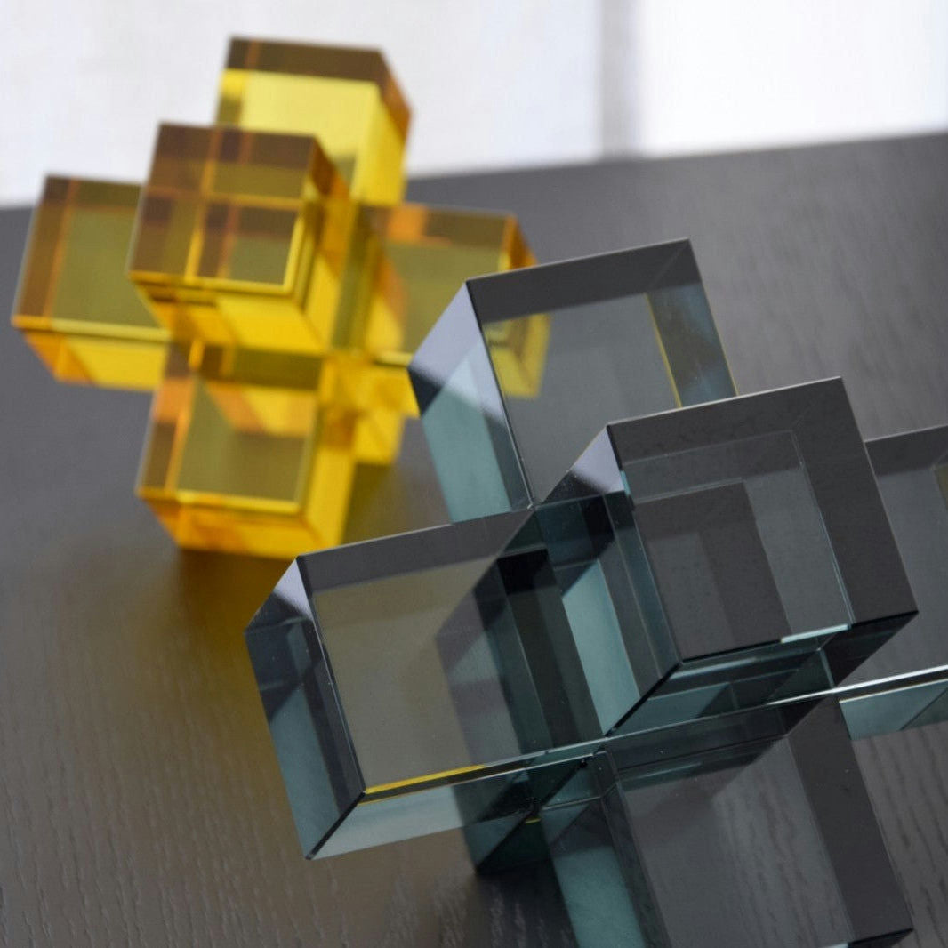 El Set de 2 Piezas Cristal Color. Cubos presenta dos cubos transparentes entrelazados, uno en amarillo vibrante y el otro en gris elegante, que descansan elegantemente sobre una superficie oscura.