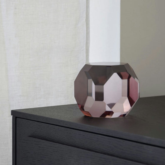 Un orbe de vidrio facetado con un tono rosado reposa sobre una superficie de madera negra cerca de una cortina blanca, agregando un toque elegante del Set de 2 Piezas en Cristal Color. Rocas a tu diseño de interiores.