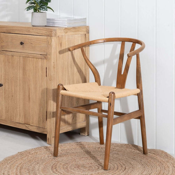 Una Silla Scandbone Marrón en Madera de Abedul con asiento tejido colocado sobre una alfombra circular junto a un armario de madera.