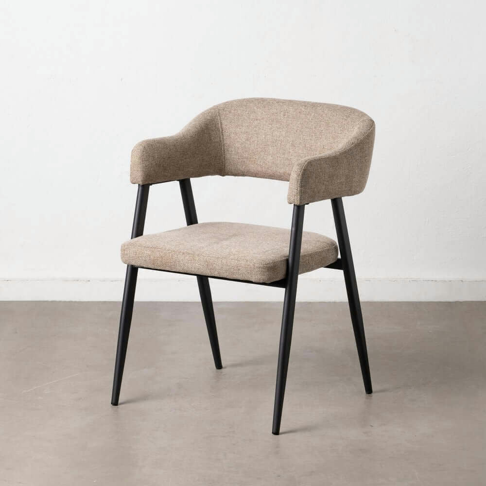 La Silla de Comedor Tapizada Topo, una silla tapizada en color beige con respaldo curvo y patas de metal negro, reposa sobre un piso de concreto contra una pared blanca, capturando un diseño moderno.