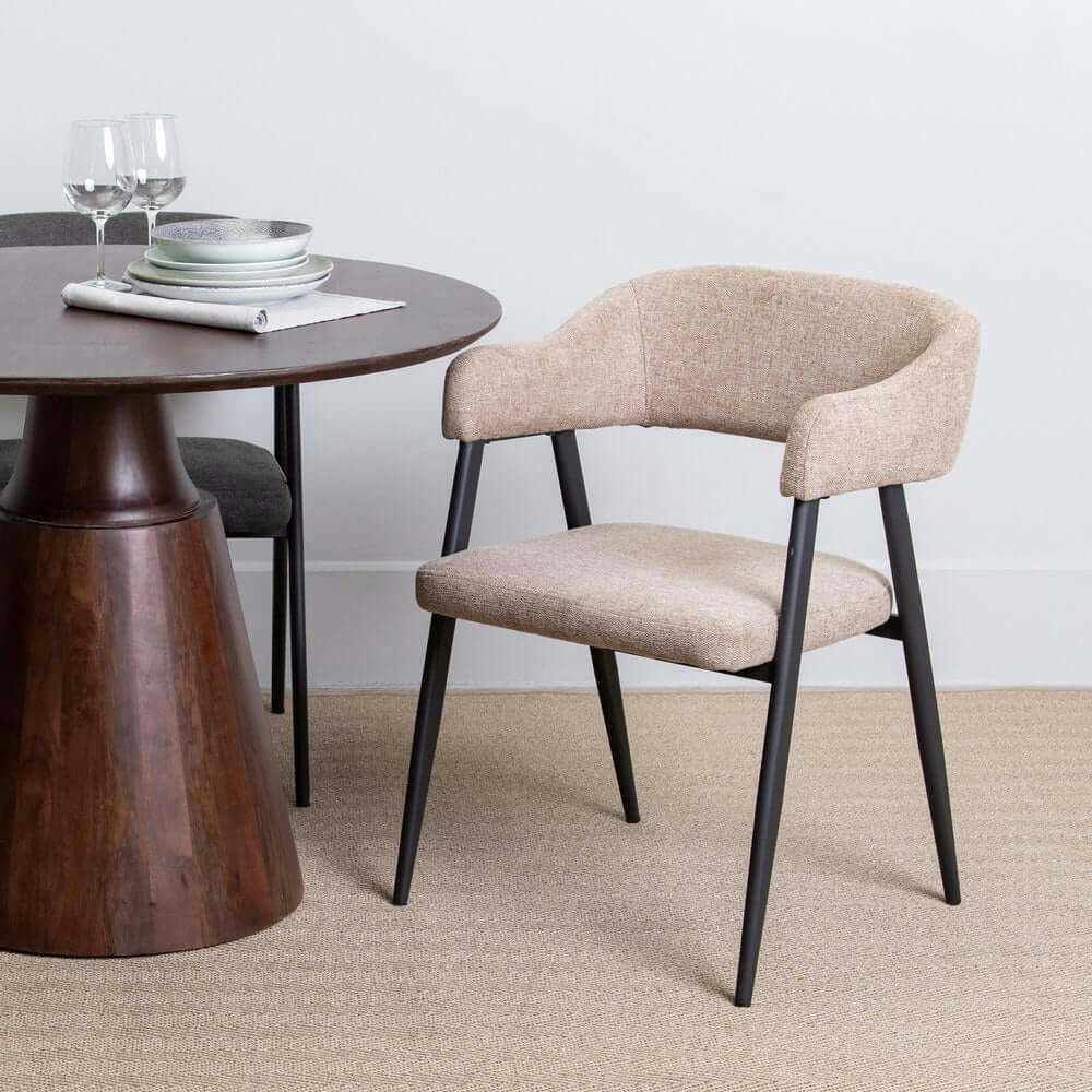 La Silla de Comedor Tapizada Topo, con elegantes patas negras y un asiento de poliéster beige, complementa perfectamente una mesa redonda de madera adornada con platos y cristalería sobre una alfombra beige.