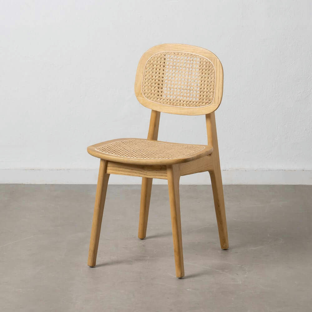 La silla de comedor de madera y ratán Nordic Elm presenta un respaldo y un asiento de ratán tejido. Ubicada sobre un piso de concreto contra una pared blanca, encarna a la perfección el estilo escandinavo.