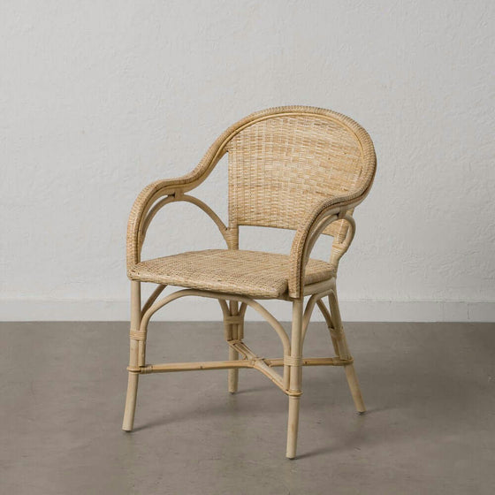 La Silla de Comedor en Ratán Natural, con respaldo curvo y apoyabrazos característicos del Estilo Costa, está elegantemente colocada sobre un piso de concreto gris contra una pared blanca lisa.