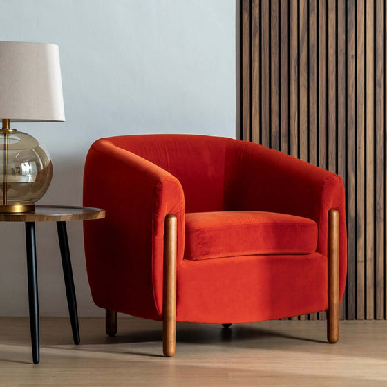 Sillón Rojo Nest de diseño moderno con patas de madera al lado de una mesa auxiliar y lámpara, contra una pared revestida de madera.