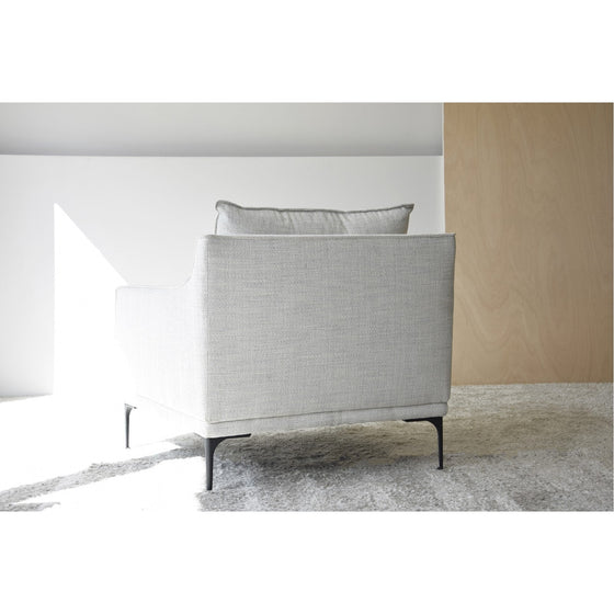 Un sillón tapizado en gris claro, que recuerda a un Sillón tapizado en Mezclilla Beige, con finas patas negras se coloca sobre una alfombra blanca junto a un panel de madera. La luz del sol resalta la parte trasera de este mueble de diseño.