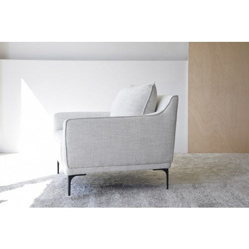 Un Sillón tapizado en Mezclilla Beige tapizado en gris claro con un cojín a juego se coloca sobre una alfombra texturizada en una habitación minimalista con paredes blancas y madera, encarnando la esencia de Mueble de diseño.