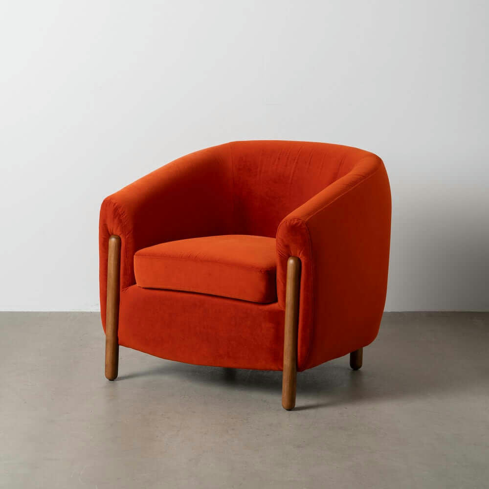 El Sillón Tapizado en Rojo Nest, con sus patas de madera, reposa elegantemente sobre una pared blanca lisa sobre un piso de concreto, mostrando un diseño moderno contemporáneo.