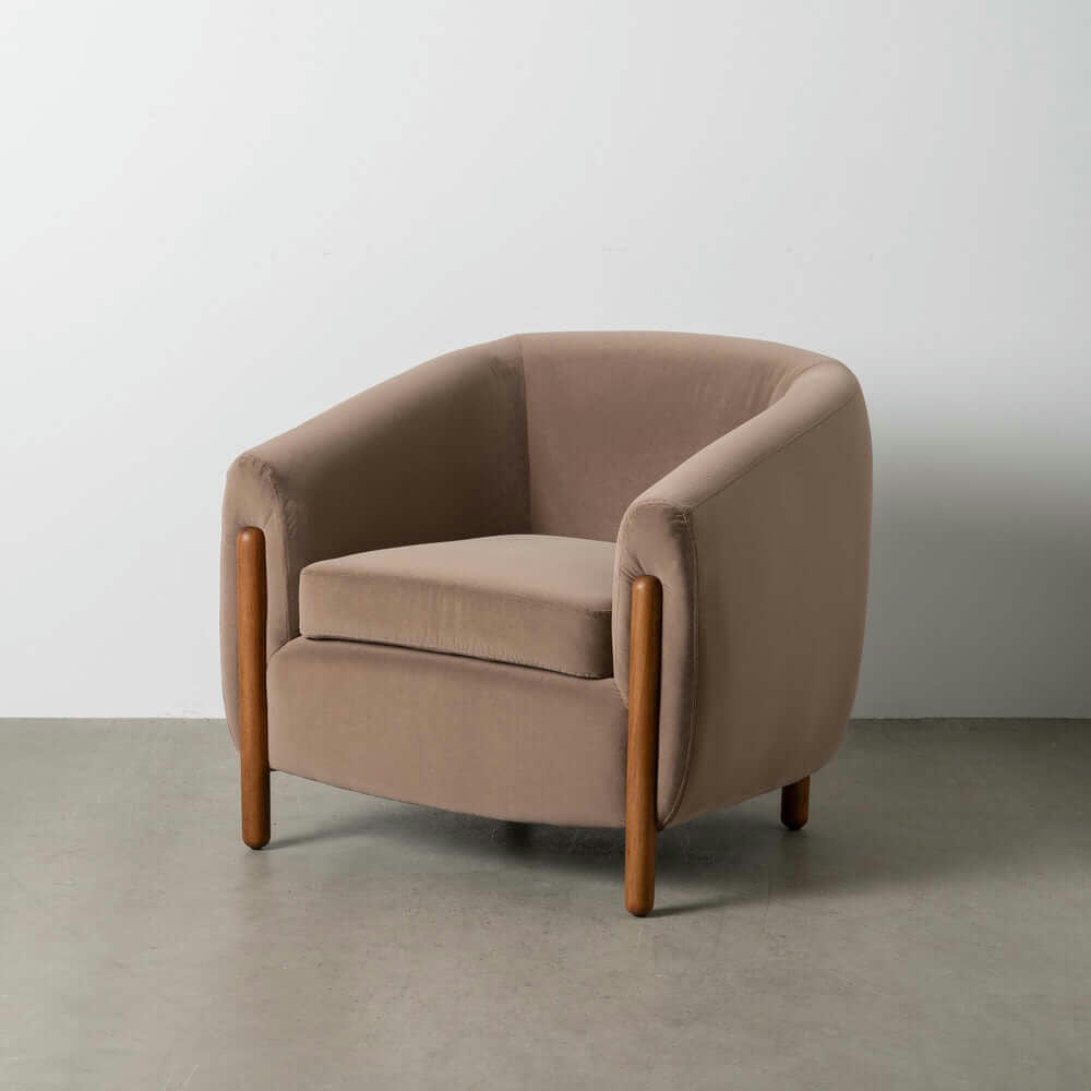 El Sillón Tapizado en Taupe Nest presenta tapizado marrón y patas de madera, ofreciendo una comodidad excepcional sobre un piso de concreto contra una pared blanca.