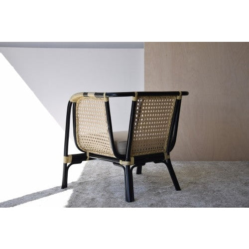 Un Sillón en Rattan con Cojín Crema negro y natural - Julia de diseño moderno se coloca sobre una alfombra de color claro en una estancia minimalista con pared de madera y luz natural, creando una decoración elegante y acorde con la decoración contemporánea.