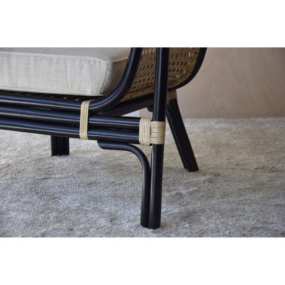 Primer plano del Sillón en Rattan con Cojín Crema - Julia con estructura de metal negro, detalles de ratán y cojín crema, sobre una alfombra texturizada de color claro, una pieza perfecta para la decoración contemporánea.