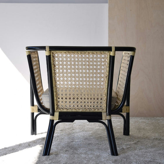 El Sillón Vintage en Ratán Julia, una silla de mimbre negra y beige con respaldo texturizado, está colocada sobre un suelo alfombrado cerca de una pared lisa, realzando la decoración contemporánea.