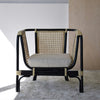 El Sillón Vintage en Ratán Julia combina el estilo vintage con una estructura de ratán y metal negro, luciendo un cojín beige sobre una alfombra gris claro, ideal para la decoración contemporánea.