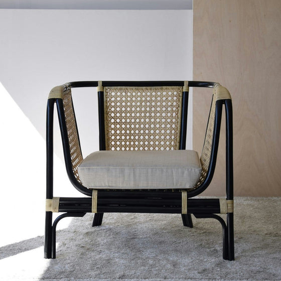 El Sillón Vintage en Ratán Julia combina el estilo vintage con una estructura de ratán y metal negro, luciendo un cojín beige sobre una alfombra gris claro, ideal para la decoración contemporánea.