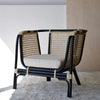 El Sillón Vintage en Ratán Julia, con marco negro y cojín beige, reposa elegantemente sobre una alfombra clara, incorporando el encanto vintage y la decoración moderna mientras complementa perfectamente el fondo de la pared de madera.