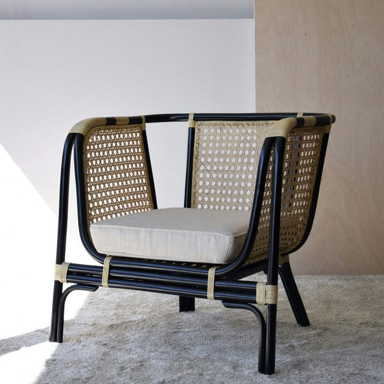 El Sillón Vintage en Ratán Julia, con marco negro y cojín beige, reposa elegantemente sobre una alfombra clara, incorporando el encanto vintage y la decoración moderna mientras complementa perfectamente el fondo de la pared de madera.
