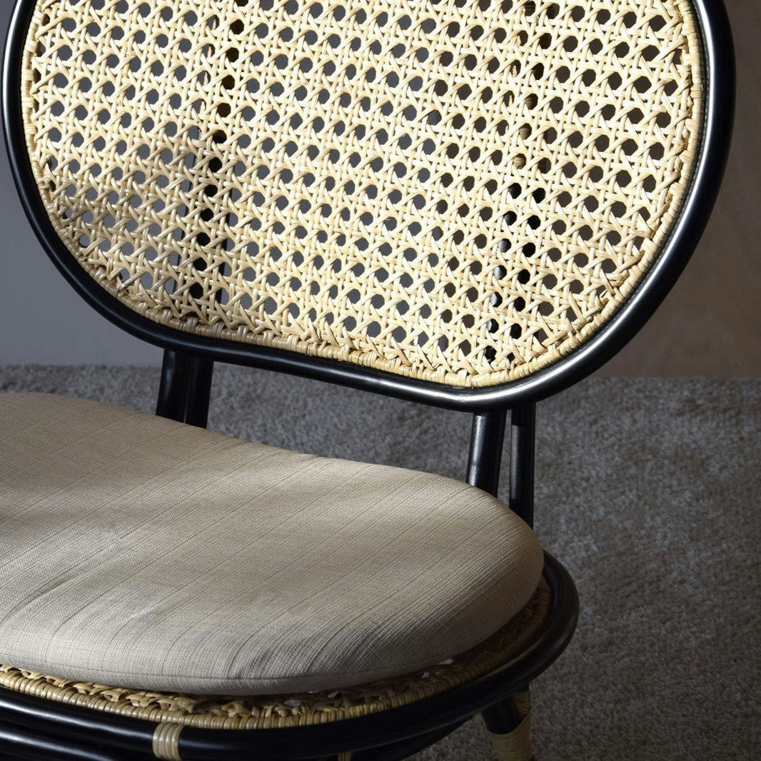 El Sillón Vintage en Ratán Norma, con marco negro y respaldo de ratán tejido, personificando el diseño exclusivo, está elegantemente colocado sobre una alfombra gris.