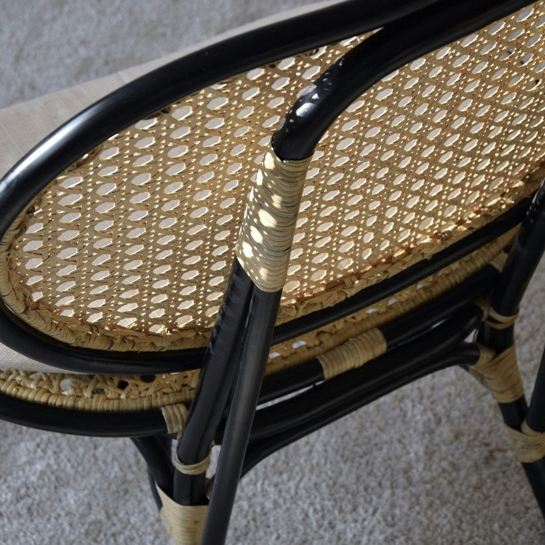 El Sillón Vintage en Ratán Norma presenta un diseño exclusivo con un marco de metal negro sobre una alfombra gris claro, aportando elegancia y encanto a cualquier espacio.