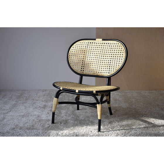 Un Sillón Vintage en Ratán Norma con estructura de metal negro y asiento y respaldo de ratán tejido, colocado sobre una alfombra gris claro contra una pared bicolor en tonos apagados de gris y madera, perfecto para decoración de interiores.