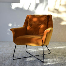  Un solo Sillón Vintage en Terciopelo Naranja "Caldero" con patas de metal negro reposa sobre una alfombra de color claro, proyectando sombras suaves en la pared.