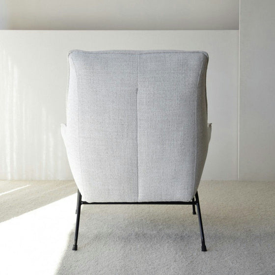 Un Sillón de Diseño Tapizado en Mezclilla "Beige" con patas de metal negro, visto desde atrás, se encuentra en una habitación luminosa y minimalista. Esta butaca de diseño complementa cualquier decoración moderna a la perfección, agregando un toque elegante a su diseño interior.